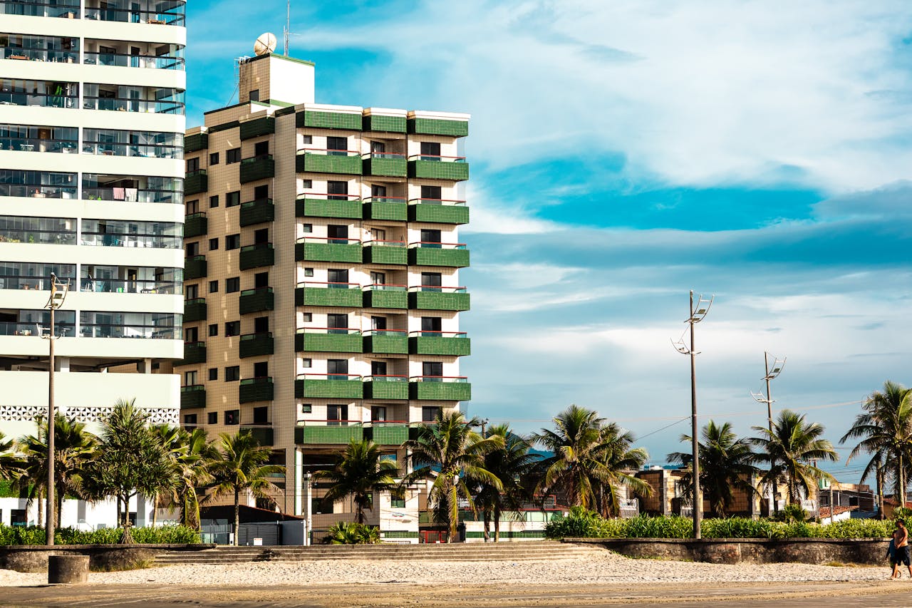 A modern apartment building next to a tropical beach with palm trees under a clear blue sky.